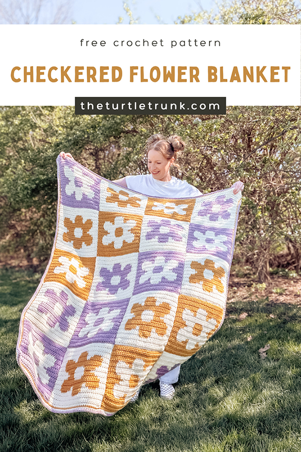 Pinterest pin for the Checkered Flower Blanket crochet pattern by The Turtle Trunk. Photo shows a woman holding up a yellow, purple, and white crochet blanket with flowers on it.