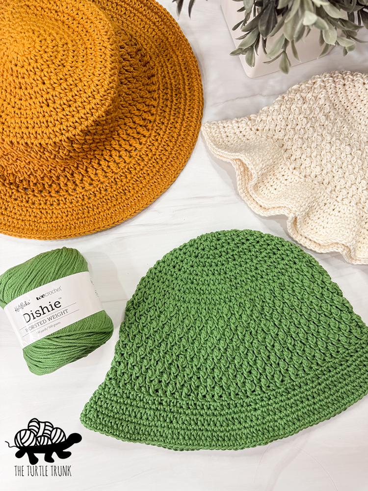 Photo shows 3 crochet sun hats laying on a white surface and a skein of green, Dishie yarn.
