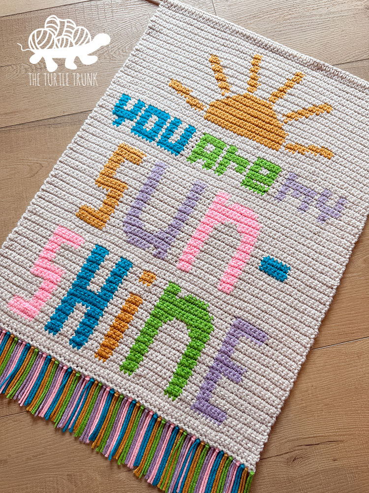 A crochet wall hanging that says You Are My Sunshine on it laying on the floor.