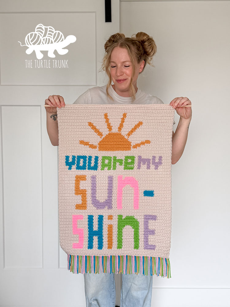 Woman holding a crochet hall hanging that says "You Are my Sunshine". Crochet Pattern designed by The Turtle Trunk.