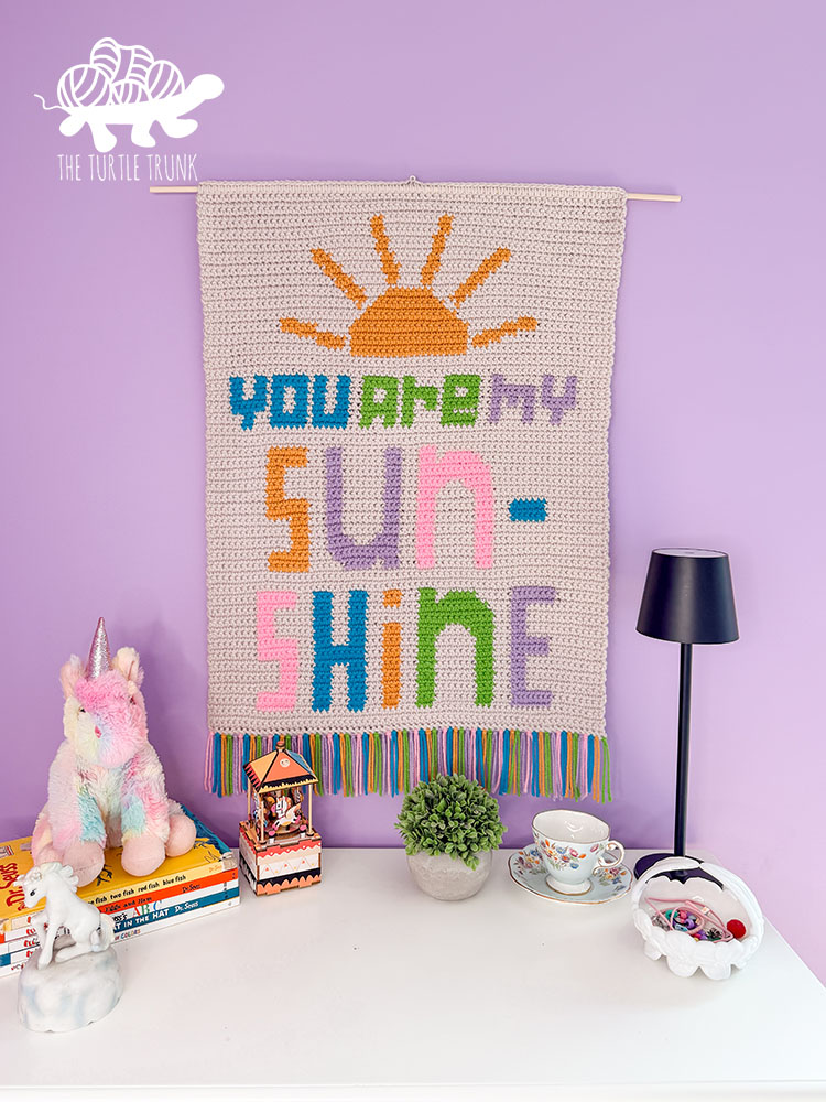 The crochet You Are My Sunshine Wall Hanging on a purple wall.