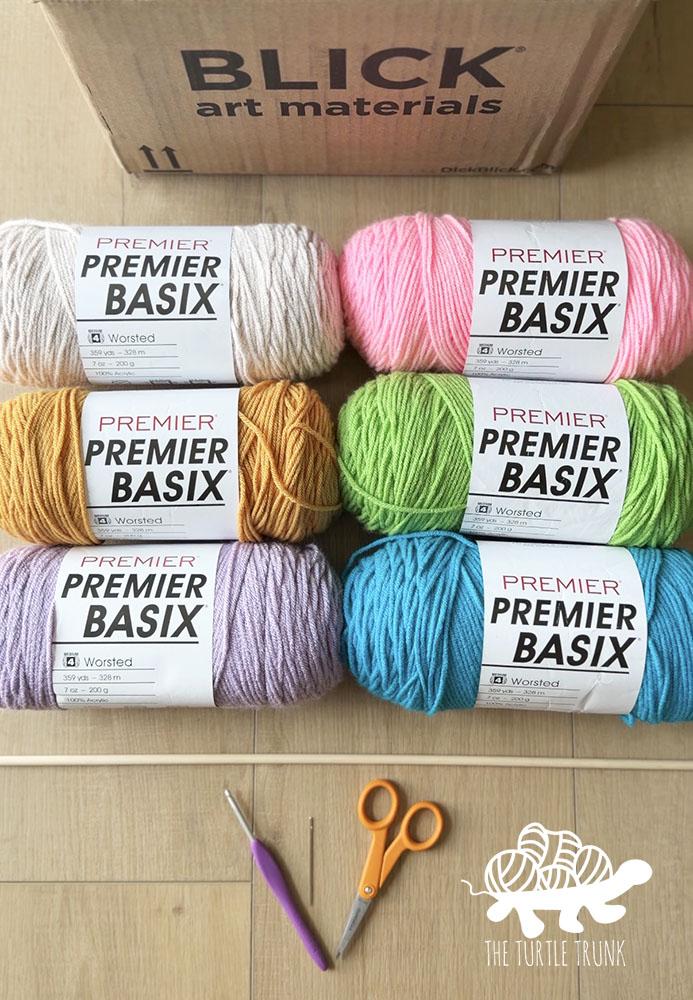 Photo shows the materials needed to make the You Are My Sunshine Wall Hanging crochet patter by The Turtle Trunk. This includes 6 skeins of yarn, a crochet hook, scissors, wooden dowel, and yarn needle.