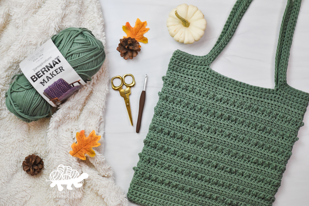 A green, crochet tote bag lays on a white surface. Picot Tote Bag crochet by The Turtle Trunk.