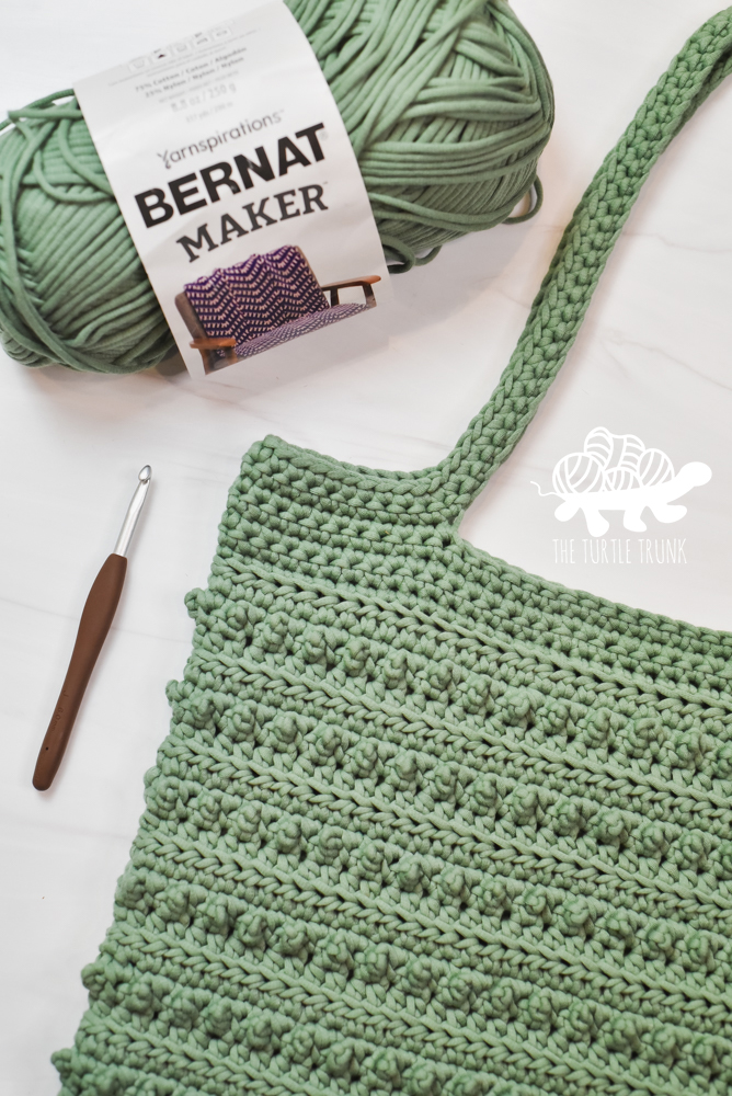 A green, crochet tote bag and a skein of Bernat Maker yarn in the color Lichen, laying on a white surface. Crochet pattern is the Picot Tote Bag by The Turtle Trunk.